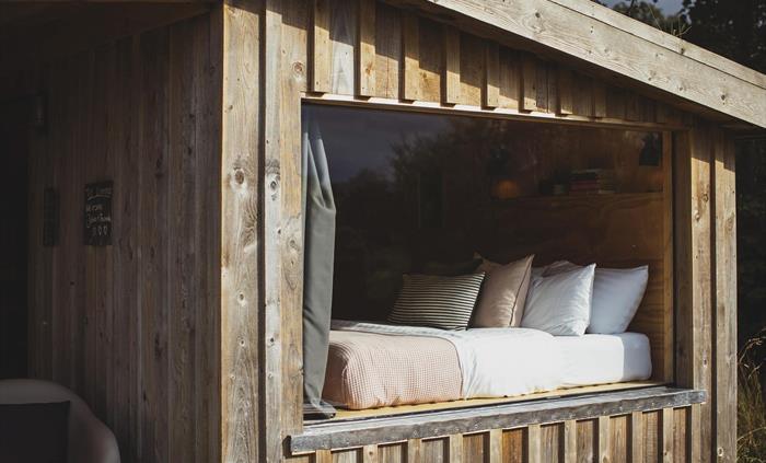 A large picture window looking into a wood cabin with a bed inside with a pink bedspread and scatter cushions.
